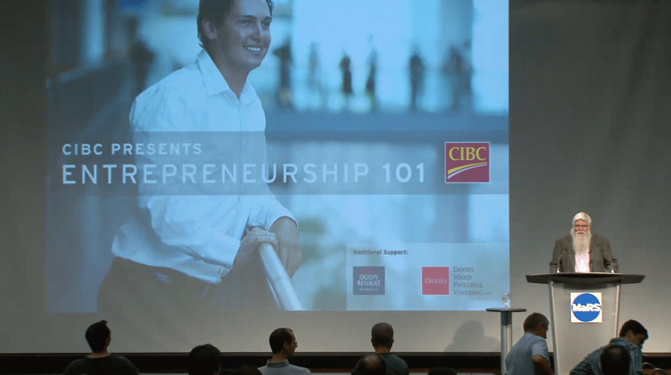 Tony Redpath speaks at the podium at an Entrepreneurship 101 lecture in 2008. An Entrepreneurship 101 image shows in the background on an overhead screen.